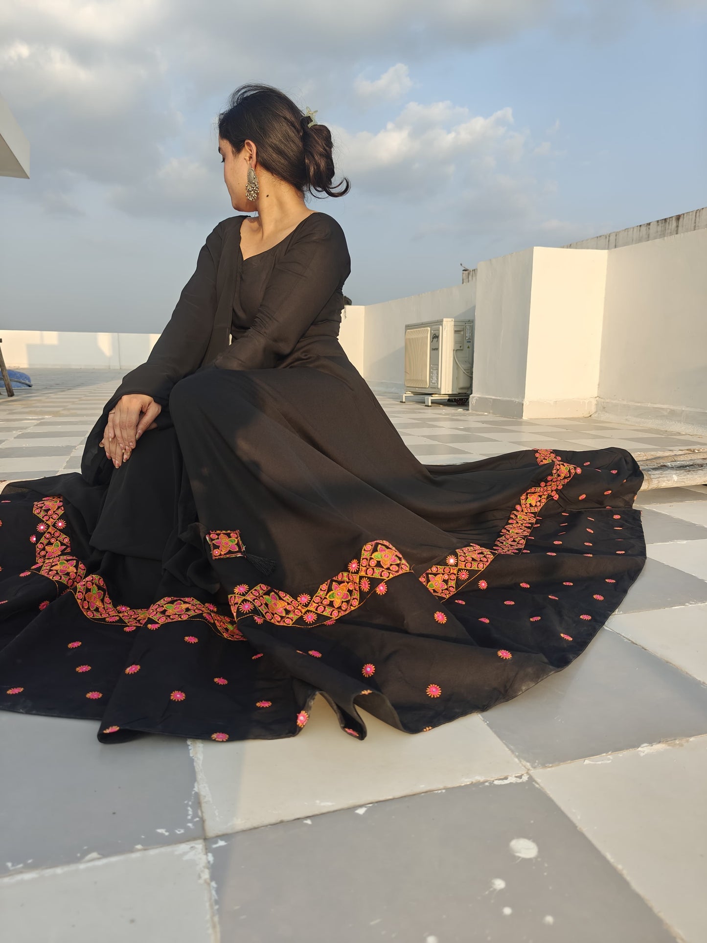 Woman in a black dress with red and orange patterns sitting on a rooftop.
