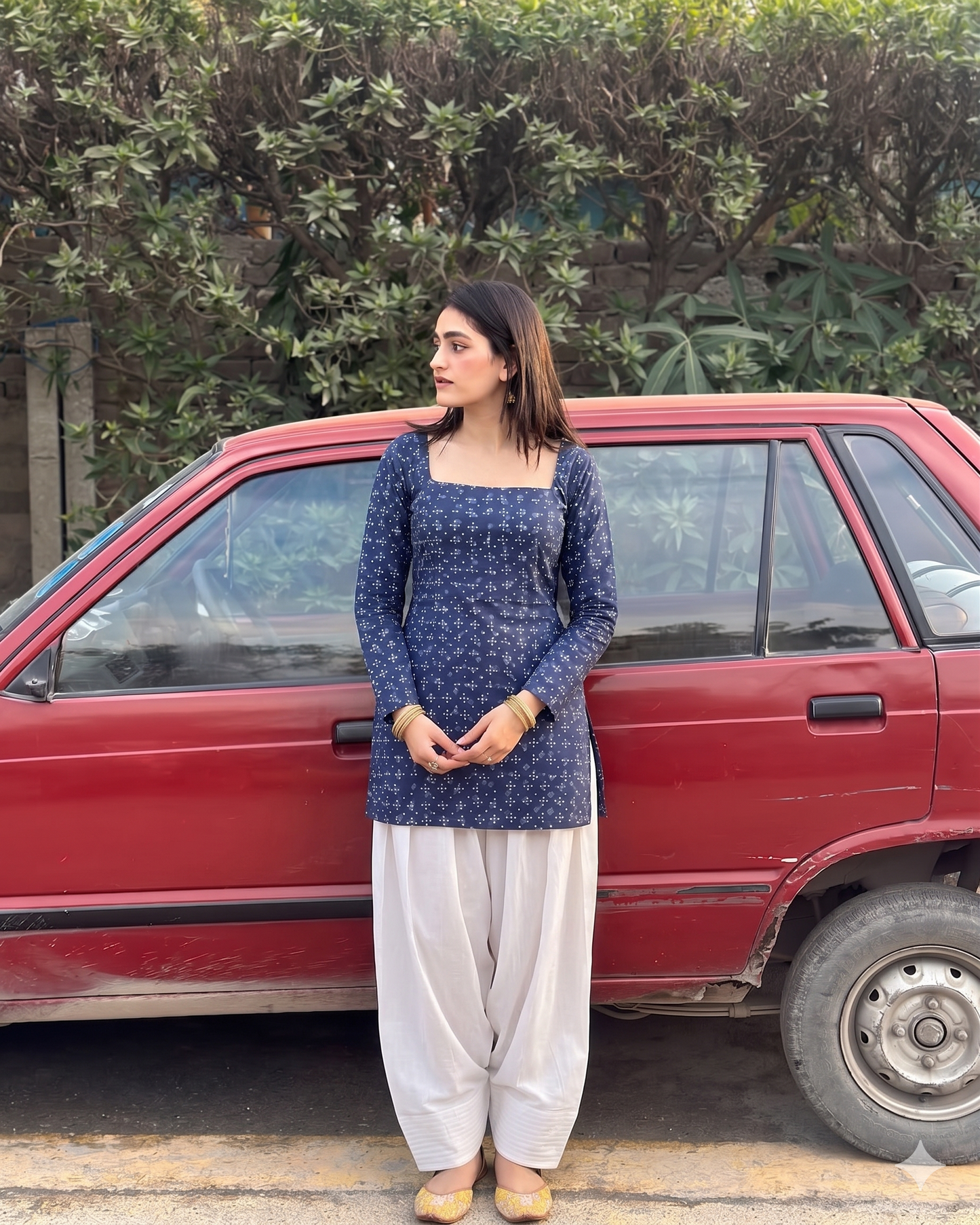 Woman in a blue top and white pants standing next to a red car with greenery in the background