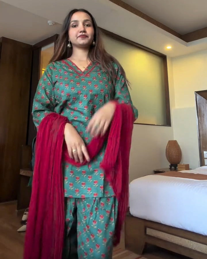 A woman modeling a green and pink patterned kurta set with a bright pink dupatta in a hotel room setting.