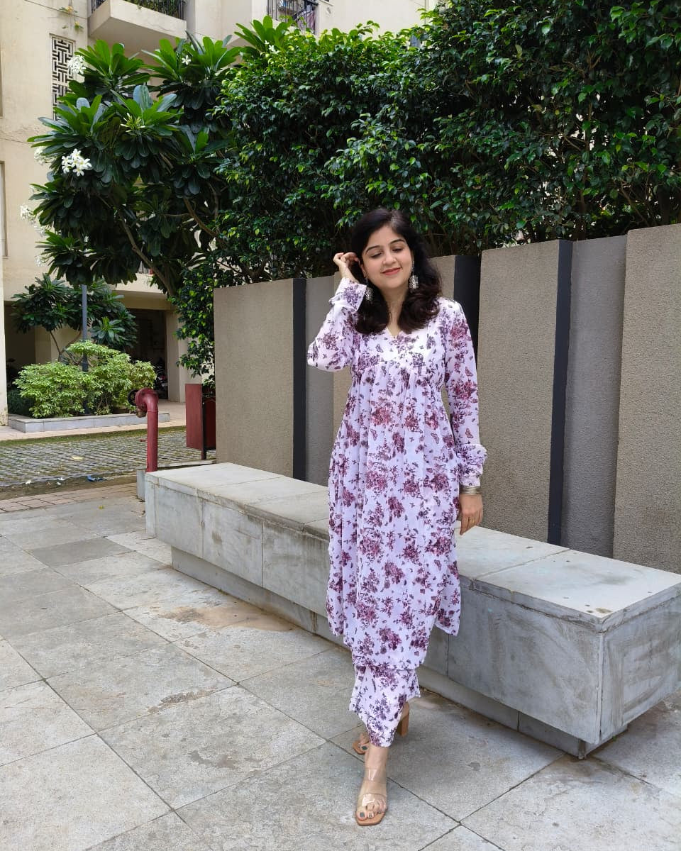 Woman in a floral dress standing outdoors near a building with greenery.