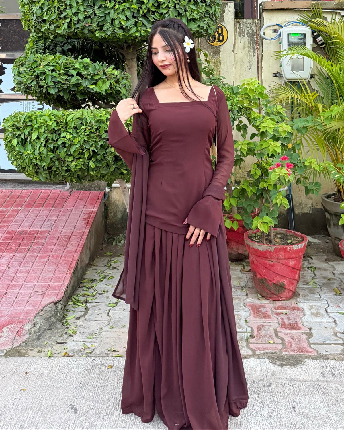 A woman wearing a long-sleeved maroon maxi dress with a matching dupatta, standing outdoors in front of a wall with potted plants.