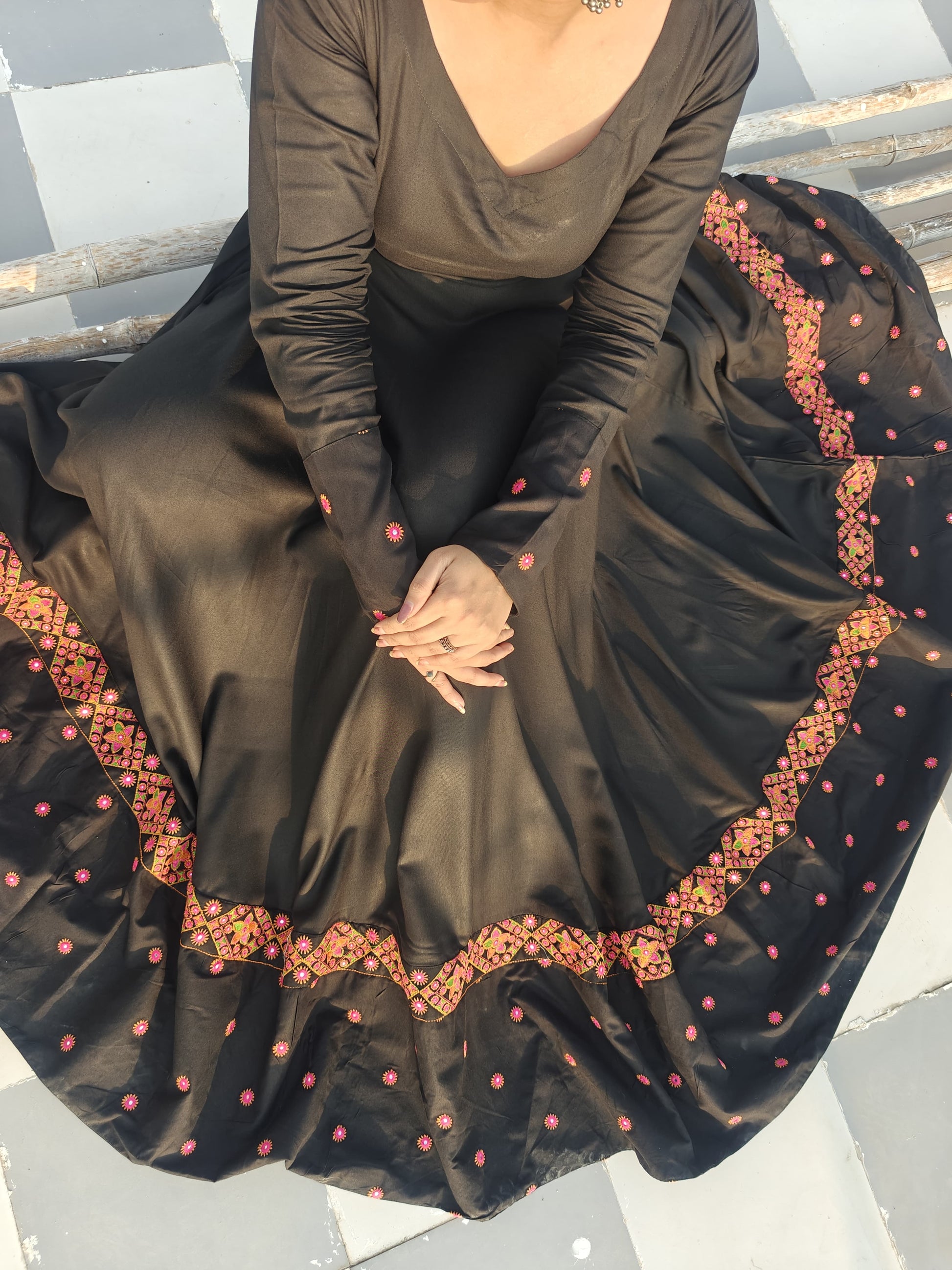 Person wearing a long black dress with intricate gold and pink embroidery sitting on  roof.