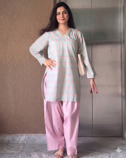 Woman wearing a light blue top with pink patterns and pink pants, standing against a neutral wall.