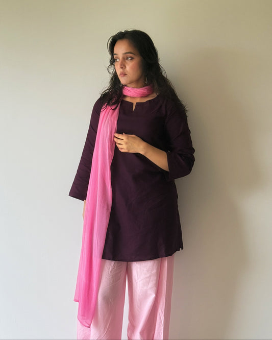 A woman modeling a traditional Indian outfit consisting of a dark plum kurta, light pink pants, and a bright pink dupatta, standing against a plain off-white wall.