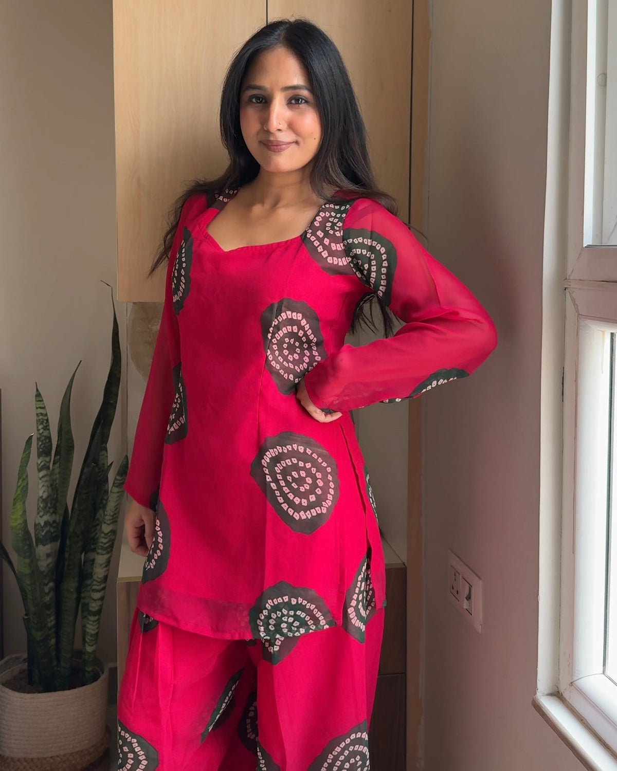 A woman modeling a bright pink traditional Indian outfit with black and green circular patterns, standing in an indoor setting next to a window.