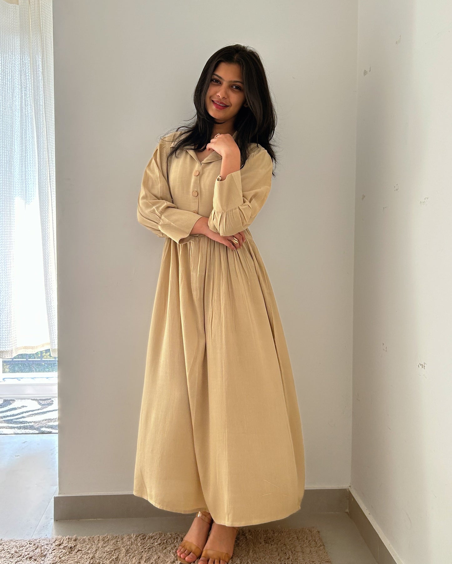 Woman wearing a beige dress standing in a minimalistic room with a window in the background.