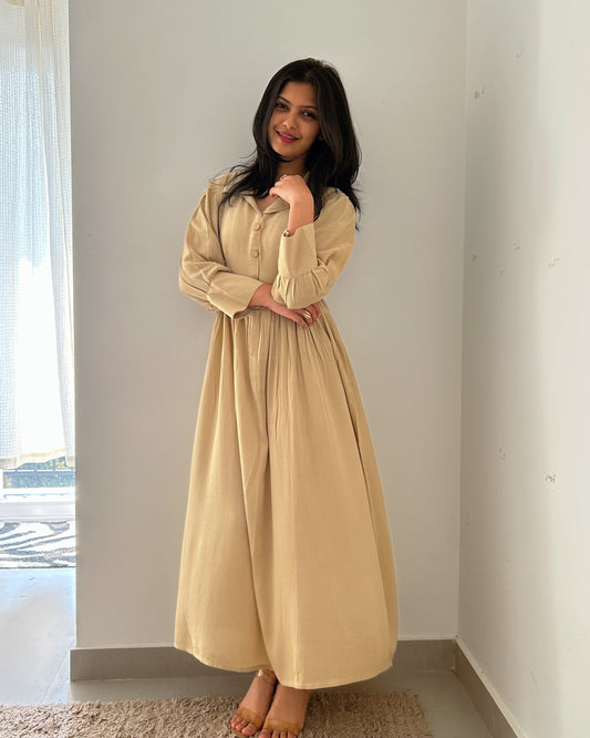 Woman wearing a beige dress standing in a minimalistic room with a window in the background.