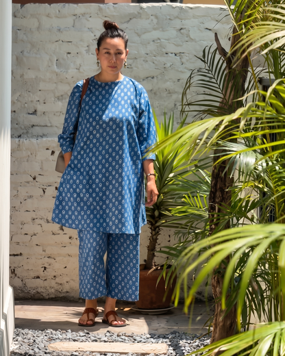 Woman in a blue patterned outfit standing on wooden steps with plants in the background