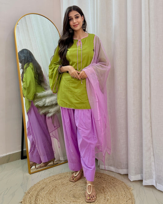 A woman models a traditional Indian outfit consisting of an olive green kurta, lavender pants, and a sheer lavender dupatta, standing in front of a mirror and white curtains.