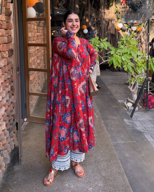 A woman stands outdoors on a walkway wearing a long red floral kurta with blue and white patterns, paired with white pants and sandals. The setting features a rustic brick wall on one side and greenery on the other.
