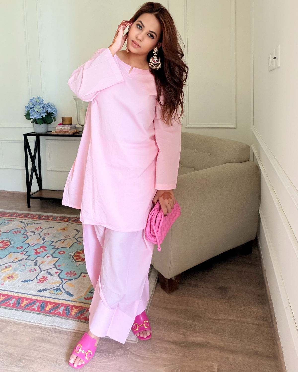 A woman modeling a light pink traditional outfit consisting of a loose long-sleeve kurta and matching trousers, holding a matching pink clutch bag in a bright, modern room with a beige sofa and patterned rug.