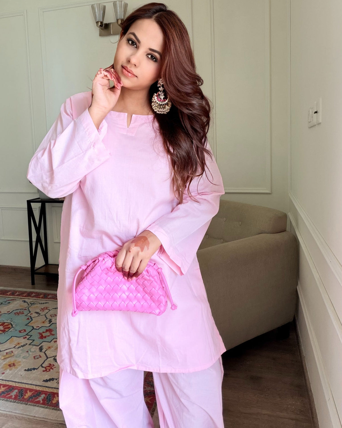 A woman wearing a light pink traditional outfit with matching pants and holding a pink quilted clutch bag, standing in a modern indoor room with white paneled walls and a beige sofa.