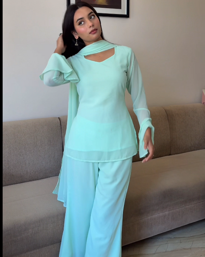 Woman in a light green outfit standing in a room with a couch and framed artwork on the wall.
