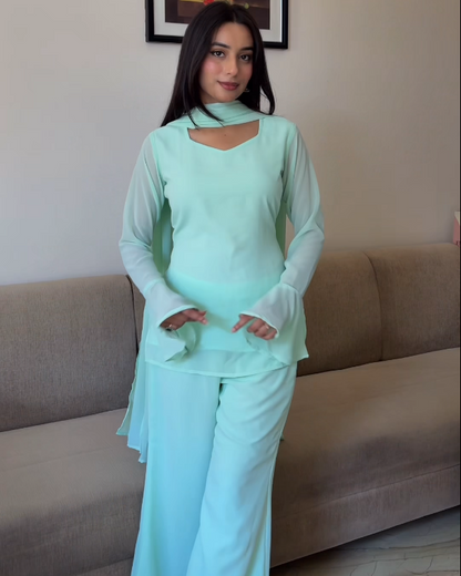 Woman in a light green outfit standing in a living room with a couch and floral painting in the background.