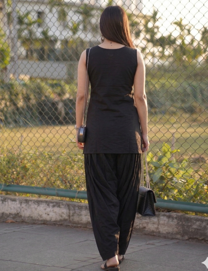 Woman in black sleeveless top and harem pants walking on a sidewalk with a chain-link fence and greenery in the background.