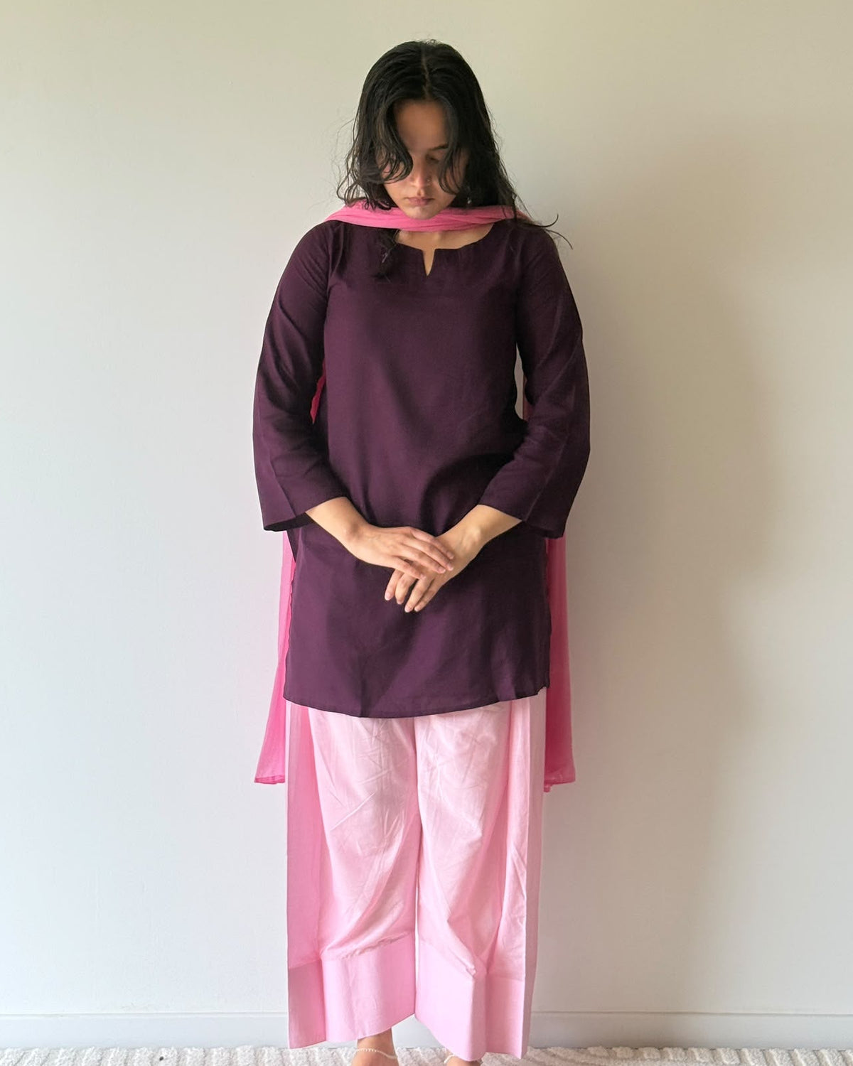 A woman stands barefoot against a plain off-white wall, wearing a dark purple tunic with a bright pink collar and pink draped fabric, paired with light pink wide-leg pants.