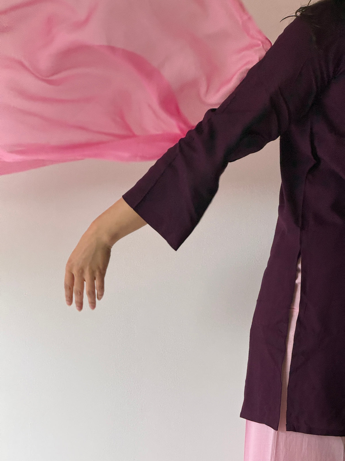 A person wearing a dark plum purple tunic with a light pink bottom and flowing pink fabric, shown from the side against a plain light background.