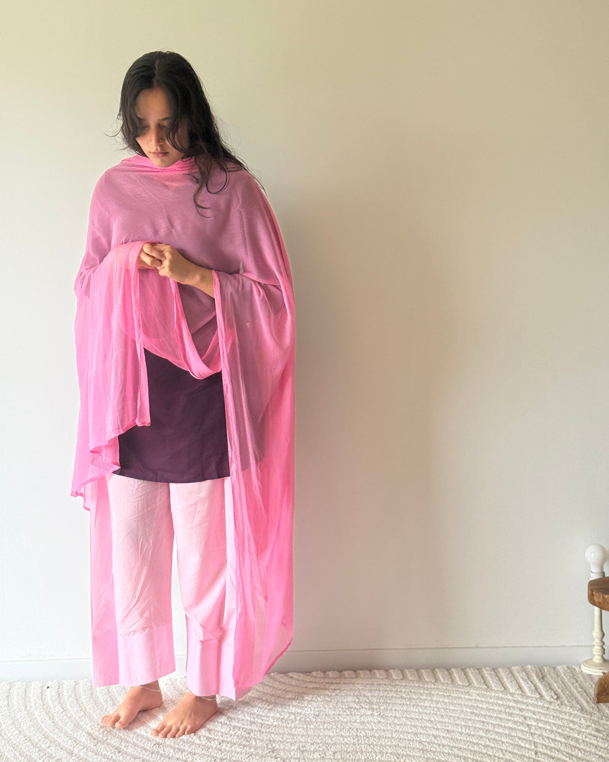 A woman stands barefoot indoors against a plain light beige wall, wearing a bright pink draped shawl or wrap over a dark purple top and light pink pants.