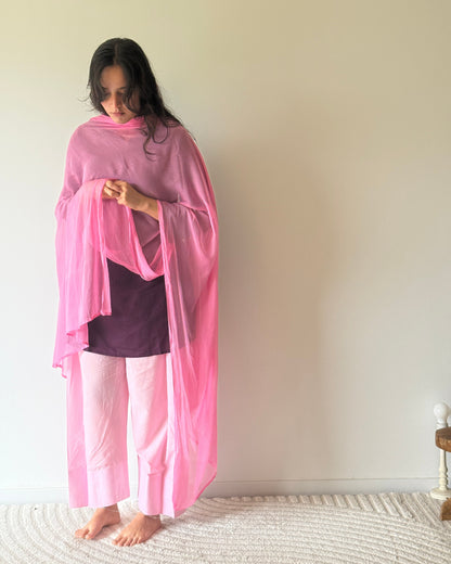 A woman stands barefoot indoors against a plain light beige wall, wearing a bright pink draped shawl or wrap over a dark purple top and light pink pants.