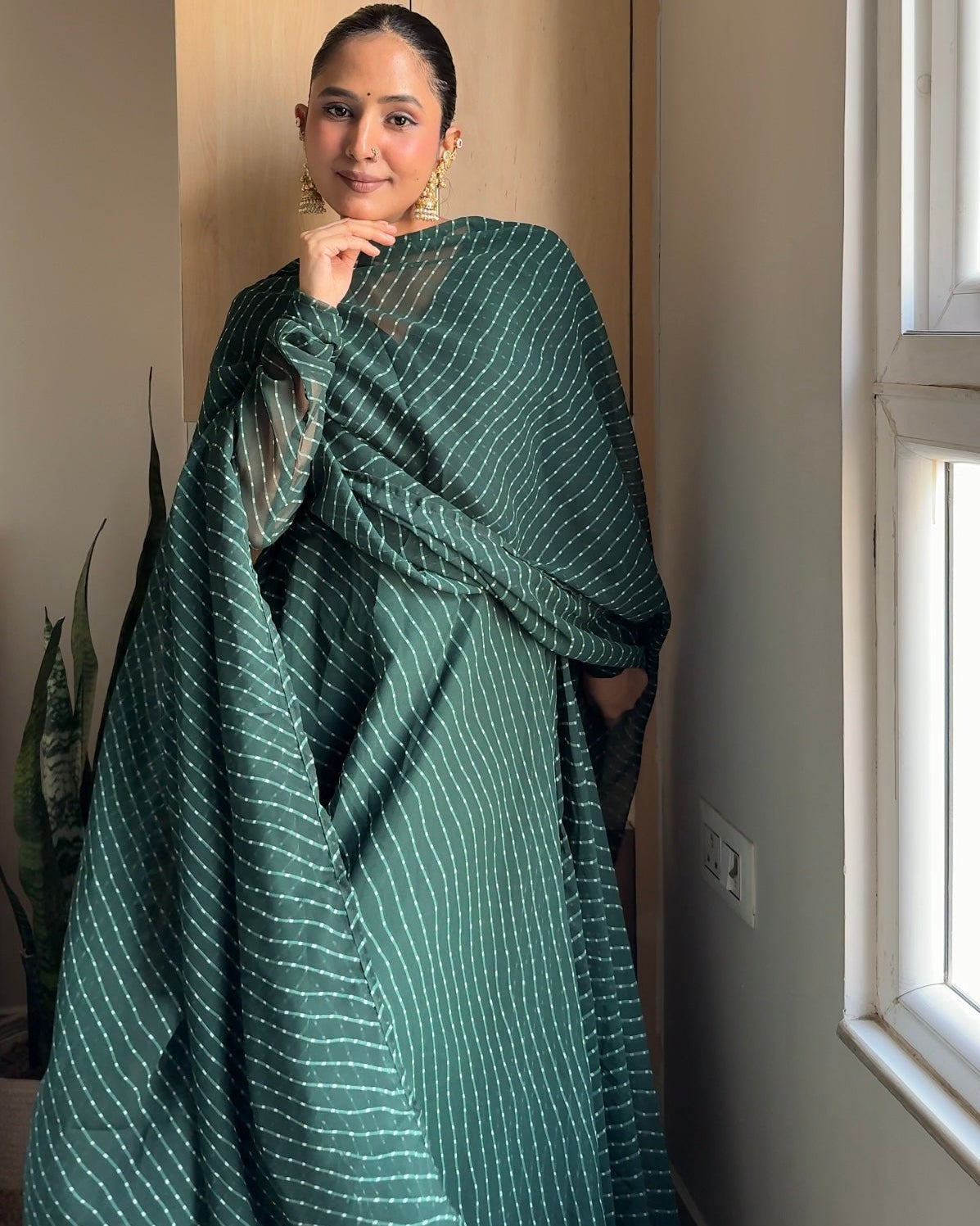 A woman wearing a dark green sari with thin white vertical stripes, standing in a brightly lit room next to a window.