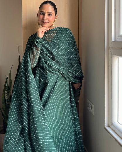 A woman wearing a dark green sari with thin white vertical stripes, standing in a brightly lit room next to a window.