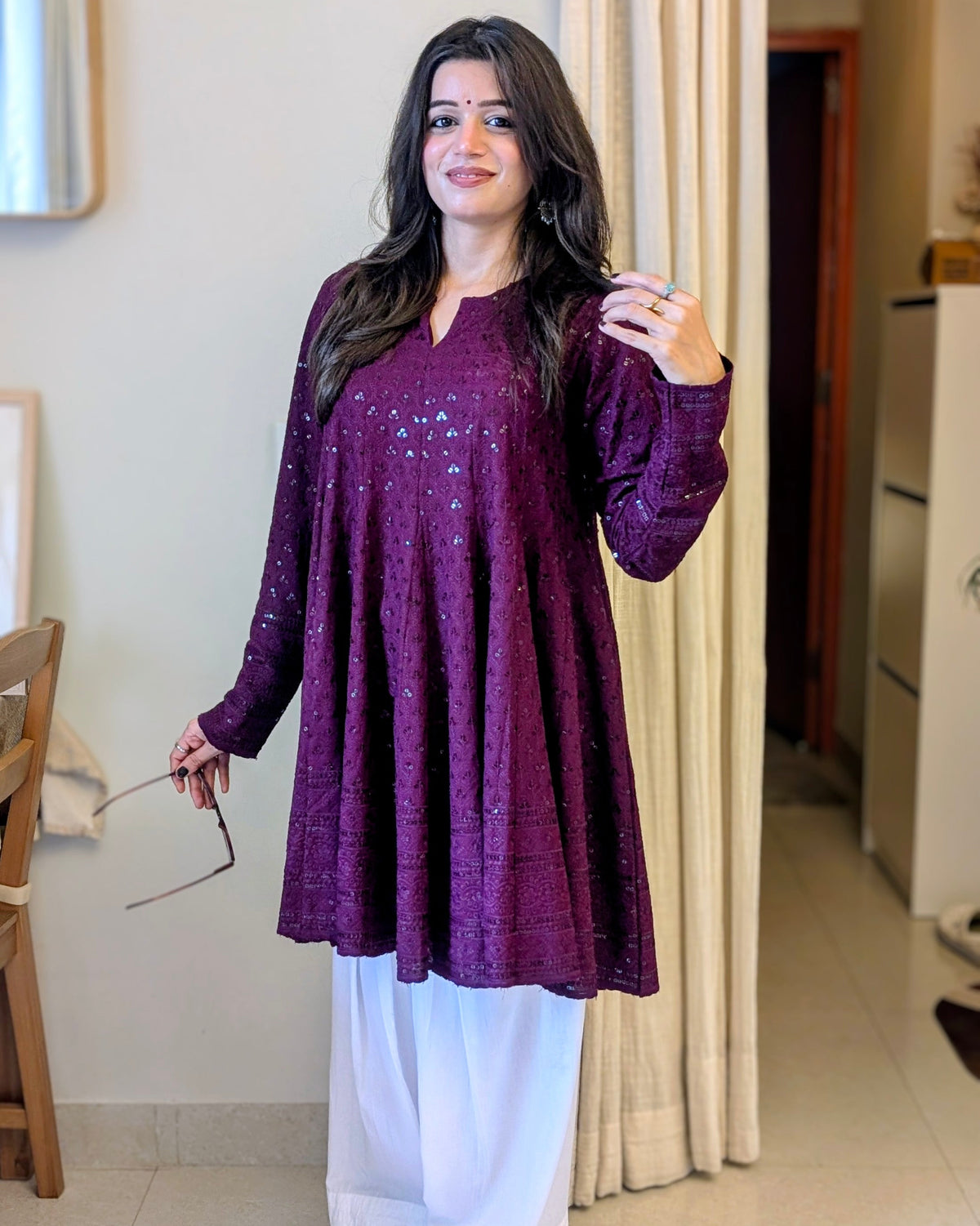 A woman modeling a deep purple long-sleeve kurta with sequin embellishments, paired with white wide-leg pants, standing in an indoor home setting.