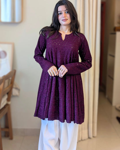 A woman wearing a long-sleeved, deep purple textured tunic top with white pants, standing in a home interior setting.