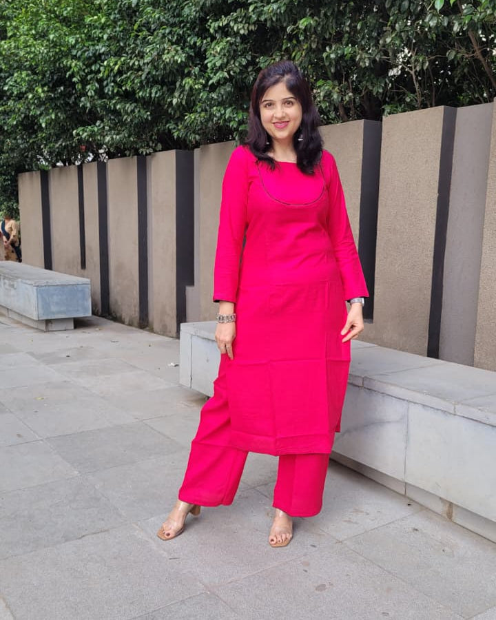 A woman wearing a bright pink kurta set with matching trousers, standing outdoors on a paved walkway in front of a modern wall and green trees.