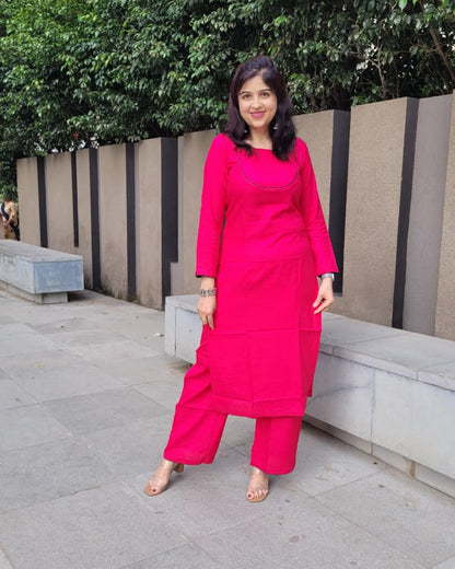 A woman wearing a bright pink kurta set with matching trousers, standing outdoors on a paved walkway in front of a modern wall and green trees.
