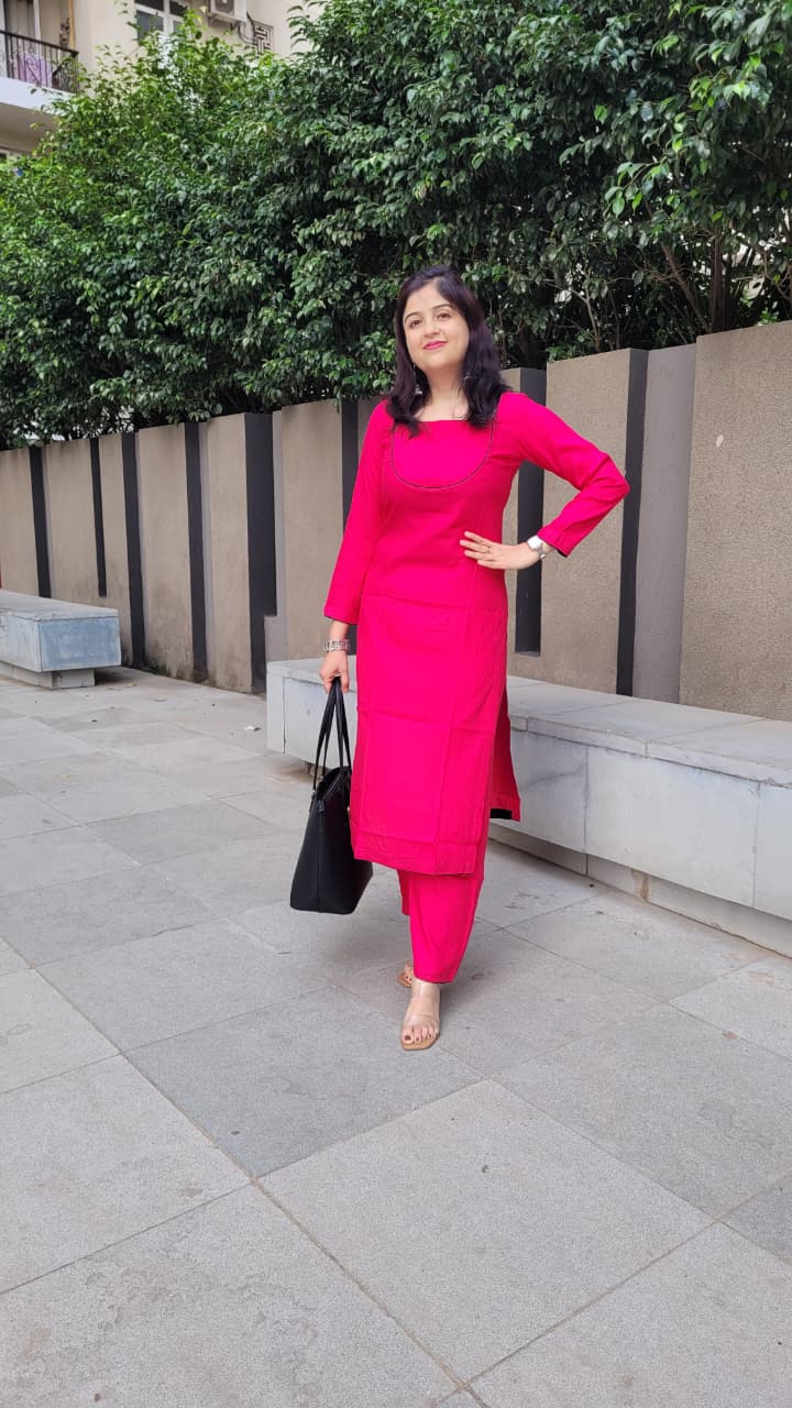 A woman wearing a bright pink kurta set with matching pants, standing outdoors on a paved walkway next to a concrete wall and green trees. She is holding a black tote bag.