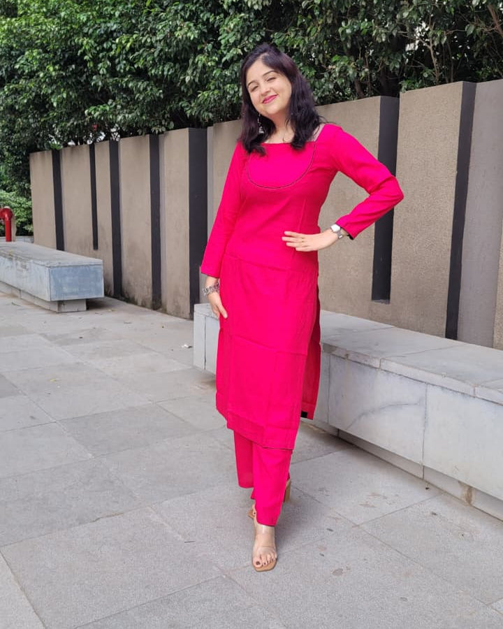 A woman wearing a bright pink long kurta and matching pants stands outdoors on a paved walkway with a modern wall and green trees in the background.