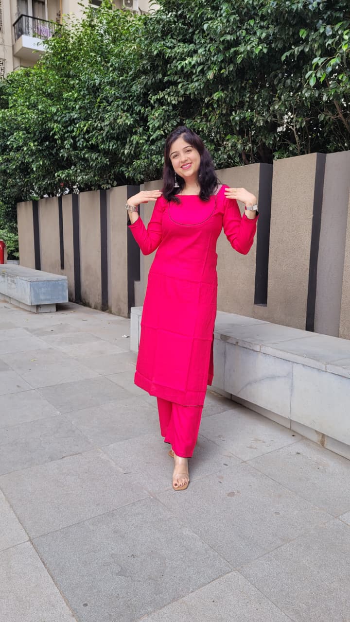 A woman wearing a bright pink kurta and matching pants stands outdoors on a paved walkway, posing with her hands on her shoulders. Trees and a modern wall are in the background.