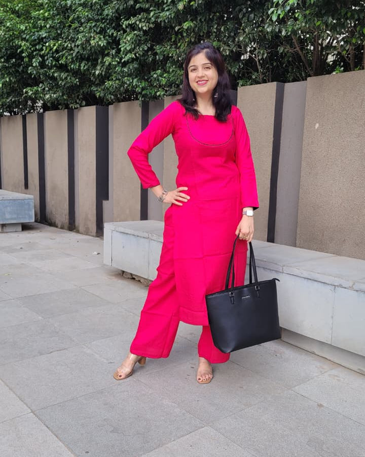 A woman posing outdoors in a bright pink kurta set, holding a black tote bag. The background features a concrete bench, green trees, and a gray tiled walkway.
