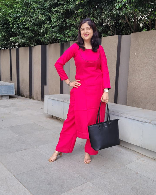 A woman posing outdoors in a bright pink kurta set, holding a black tote bag. The background features a concrete bench, green trees, and a gray tiled walkway.