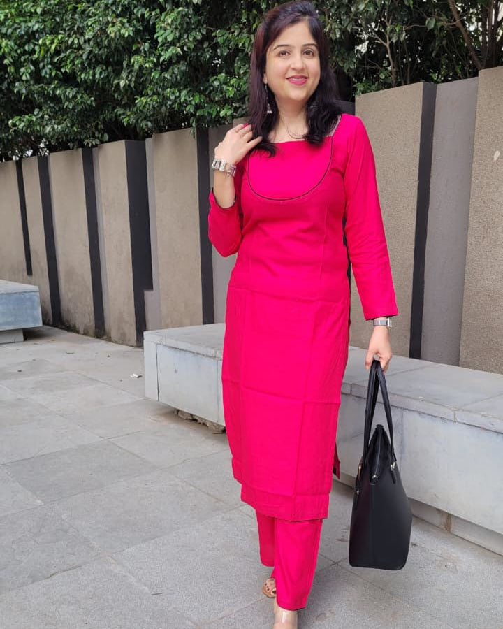 A woman wearing a bright pink kurta and matching trousers, holding a black handbag, standing outdoors on a paved walkway.