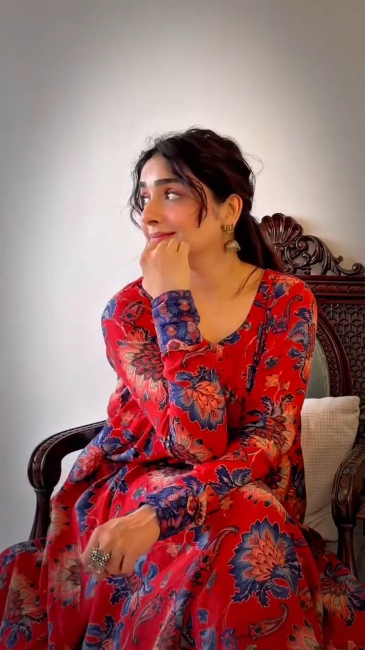 A woman wearing a red long-sleeve floral dress sits on a dark wooden chair against a plain light gray wall.