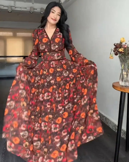 A woman wearing a long-sleeved, floor-length floral dress with a V-neck and waist tie, posing in an indoor room with a chandelier and track lighting.