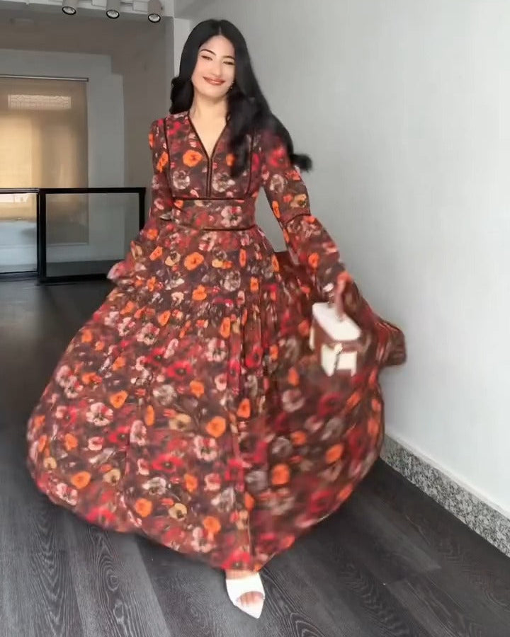 A woman wearing a long-sleeved, floor-length floral dress with a brown base and orange, red, and yellow floral pattern, captured in a wide twirl pose inside a modern room with white walls and dark wood floors.