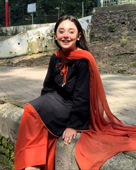 A young woman smiling while sitting outdoors on a stone ledge, wearing a black traditional outfit with a bright rust-orange dupatta draped over her shoulders.