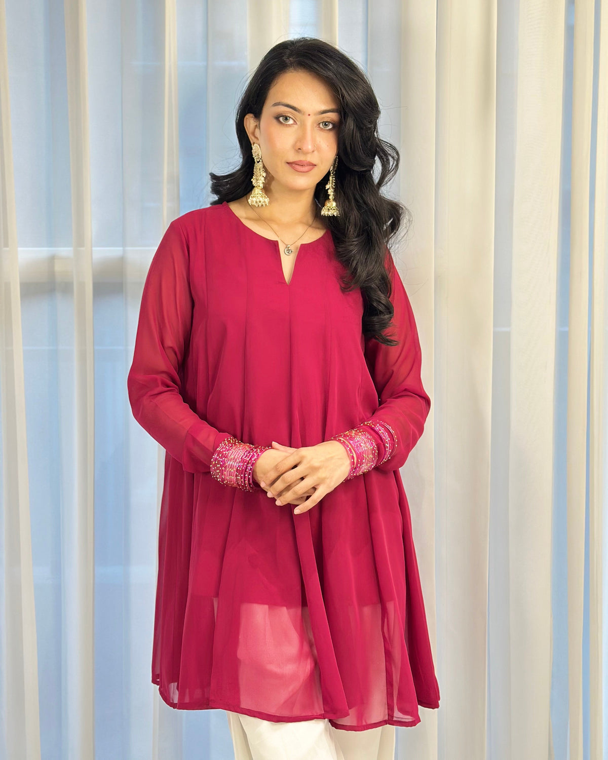 A woman modeling a long-sleeve magenta pink tunic with embroidered cuffs, paired with white pants, standing in front of a light-colored curtain backdrop.