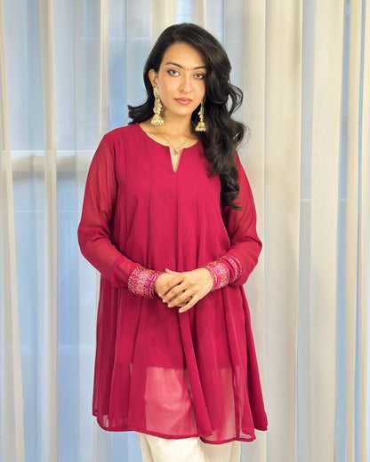 A woman modeling a long-sleeve magenta pink tunic with embroidered cuffs, paired with white pants, standing in front of a light-colored curtain backdrop.