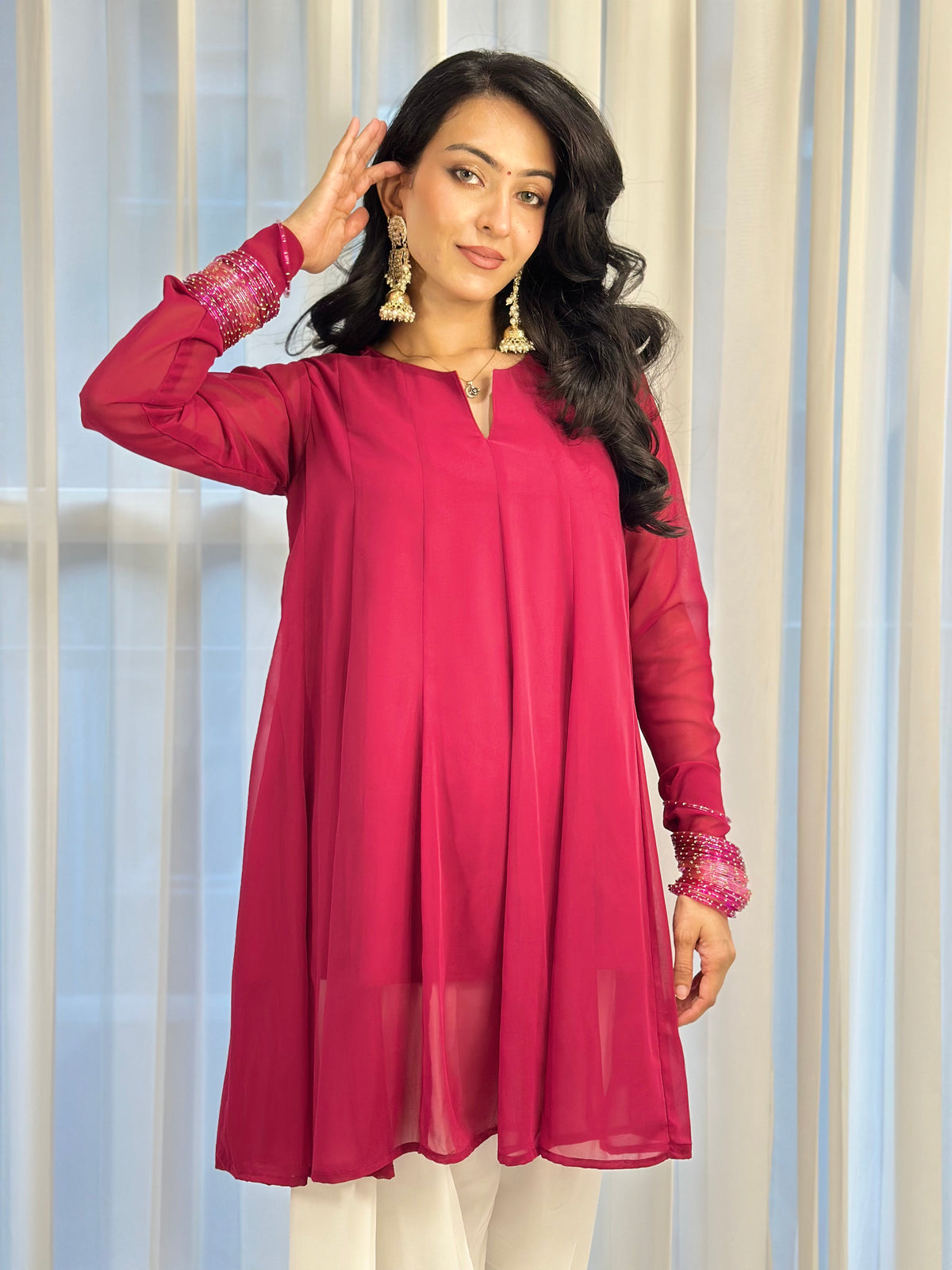 A woman modeling a bright magenta pink long-sleeve kurta with sheer sleeves and embroidered cuffs, paired with white pants, standing in front of a light-colored curtain background.