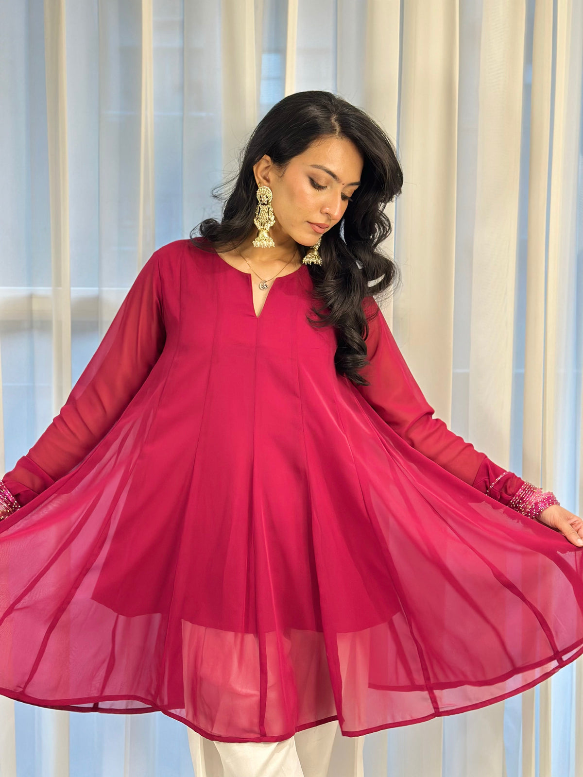 A woman wearing a bright magenta pink sheer long-sleeve kurta with white pants, standing in front of white curtains and holding out the sides of the skirt to show its flow. She is wearing gold earrings.