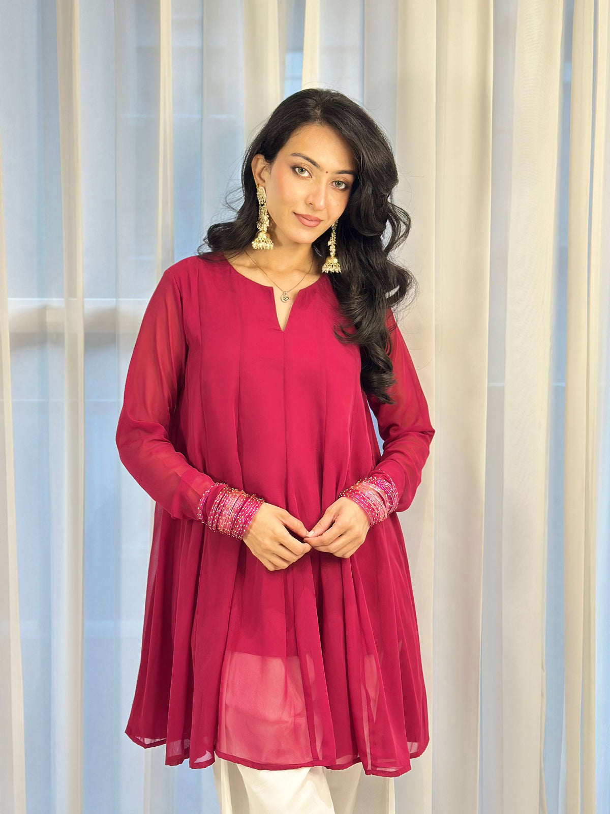A woman modeling a magenta pink long-sleeve kurta with embroidered cuffs, paired with white trousers, standing in front of white sheer curtains.