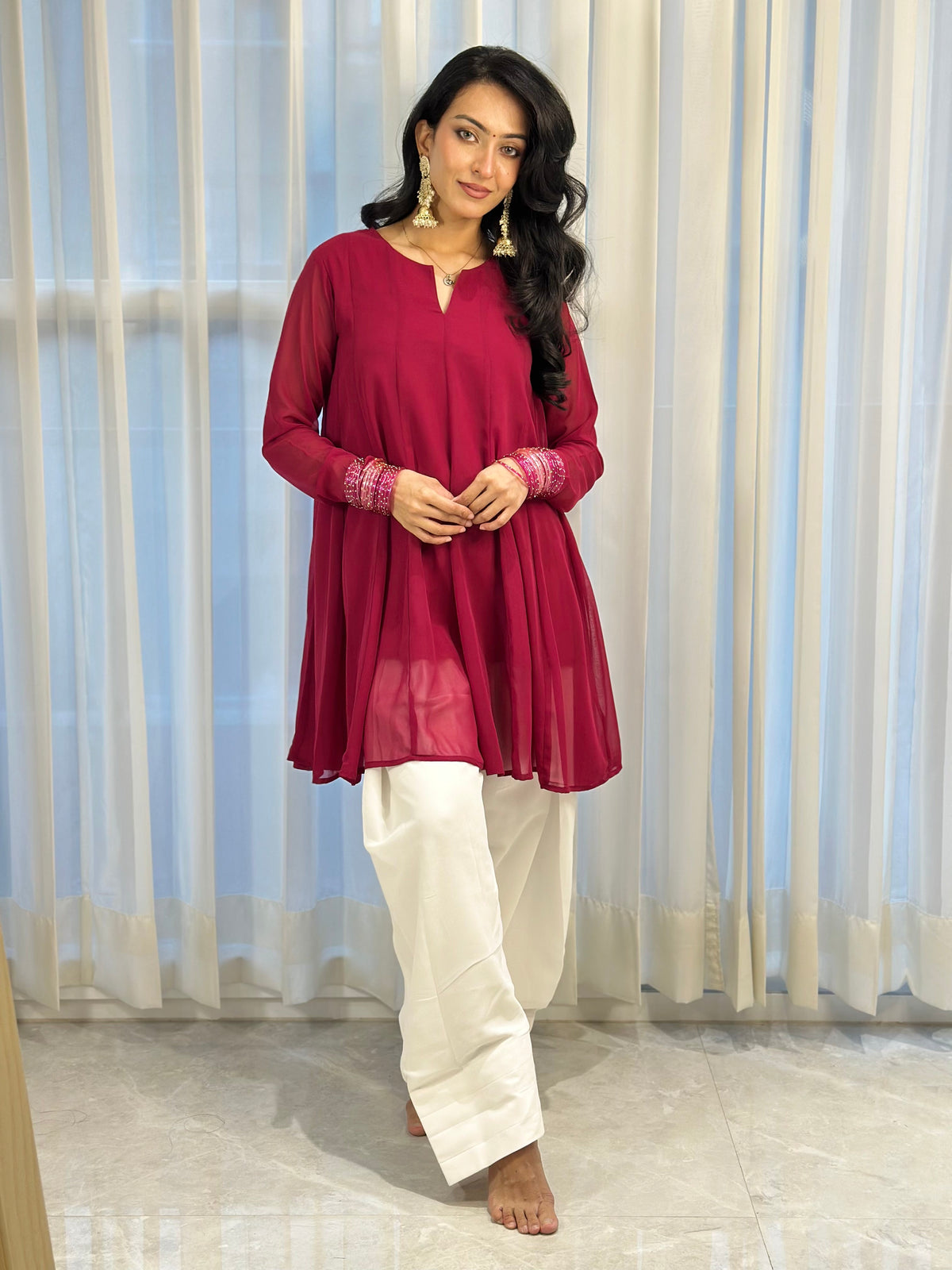 A woman modeling a long-sleeve magenta tunic top with pink embroidered cuffs, paired with white wide-leg pants, standing in front of white curtains.