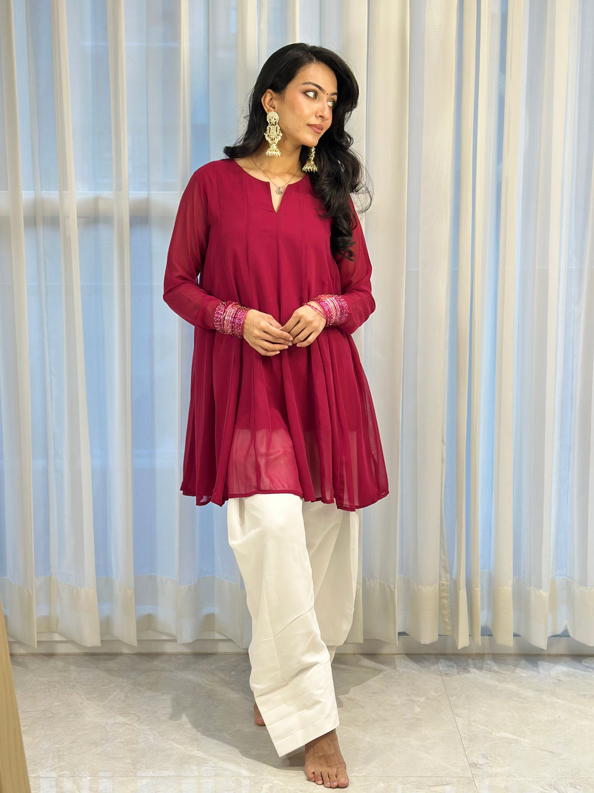 A woman modeling a deep magenta long-sleeve kurta with embroidered cuffs, paired with white wide-leg trousers, standing in front of white sheer curtains.