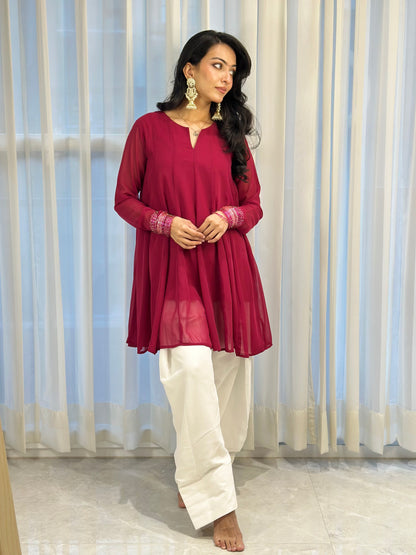 A woman modeling a deep magenta long-sleeve kurta with embroidered cuffs, paired with white wide-leg trousers, standing in front of white sheer curtains.