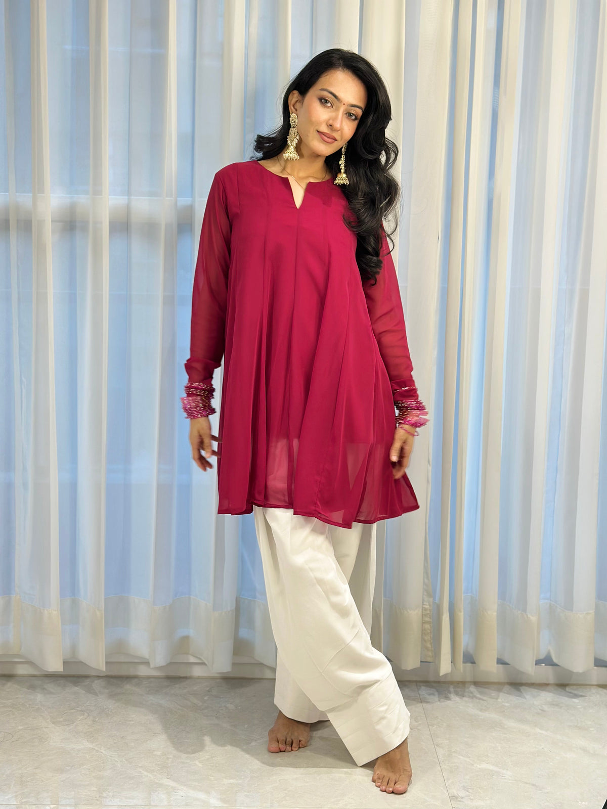 A woman modeling a magenta long-sleeve kurta with white wide-leg pants, standing barefoot in front of white sheer curtains.