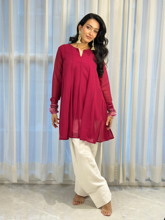 A woman modeling a magenta long-sleeve kurta with white wide-leg pants, standing barefoot in front of white sheer curtains.
