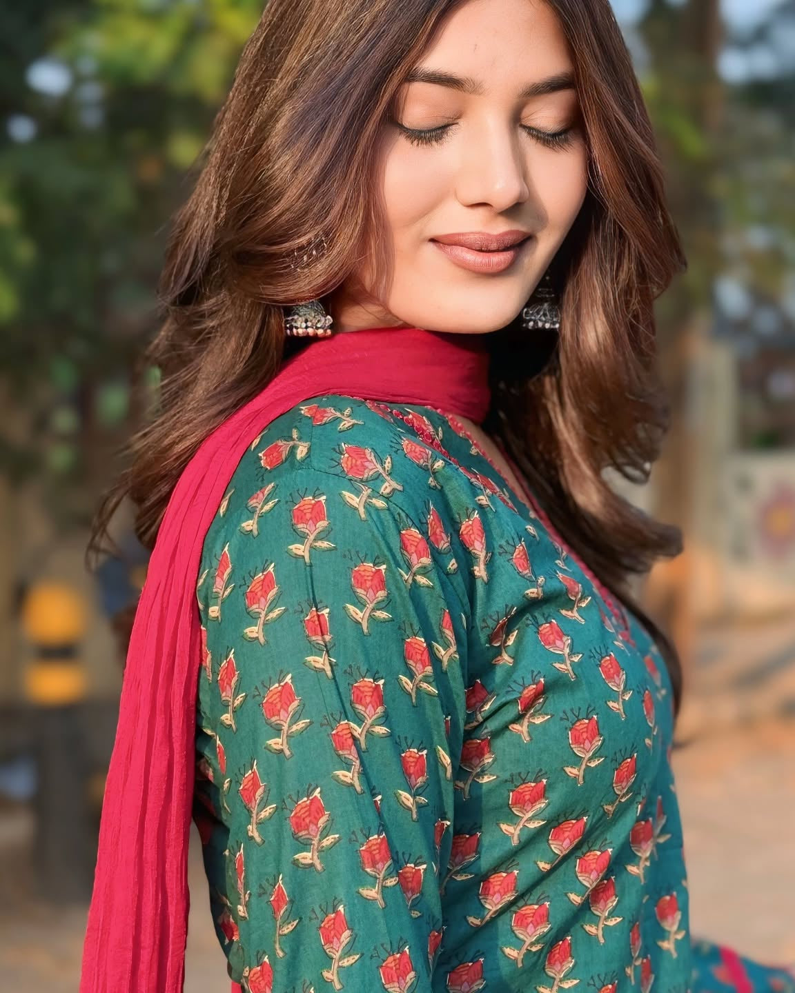 A woman wearing a green salwar kameez suit with a red floral print and a bright pink dupatta, photographed outdoors with a blurred background.
